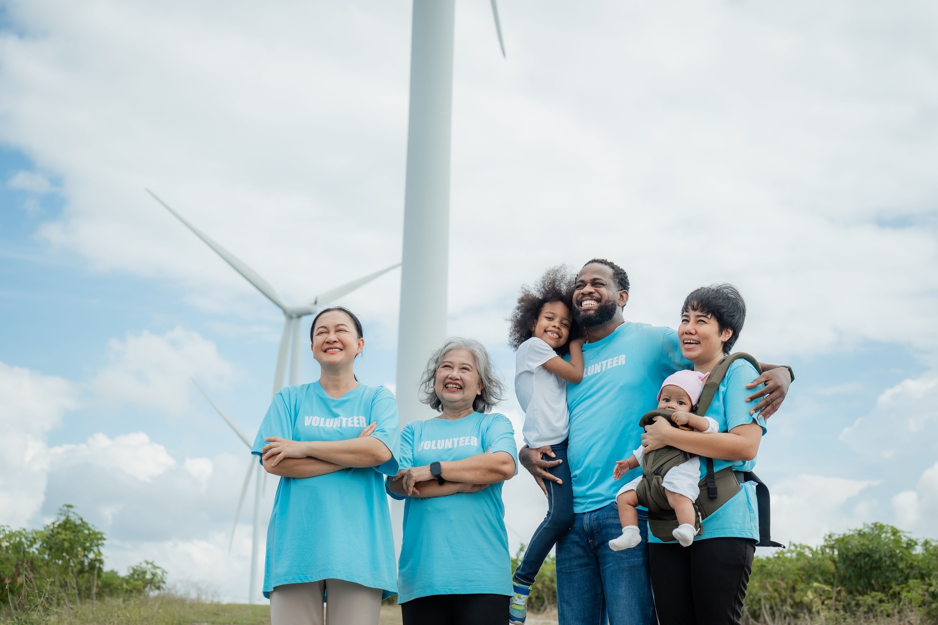 A group of people wearing blue shirts with the word "infinity" on them are standing in front of a wind turbine. environment, clean, wind turbine, volunteer, community, teamwork, social, care, nature